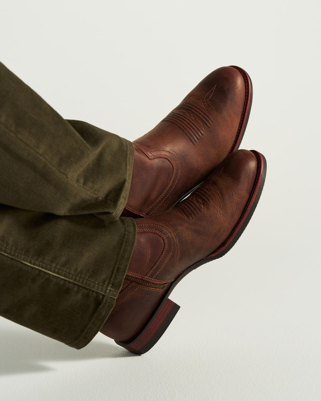 The Wayne in Maplewood Cowhide. Person wearing olive green pants and brown leather cowboy boots, featuring rich Maplewood Cowhide, shown from the side with feet crossed and resting on a light surface.