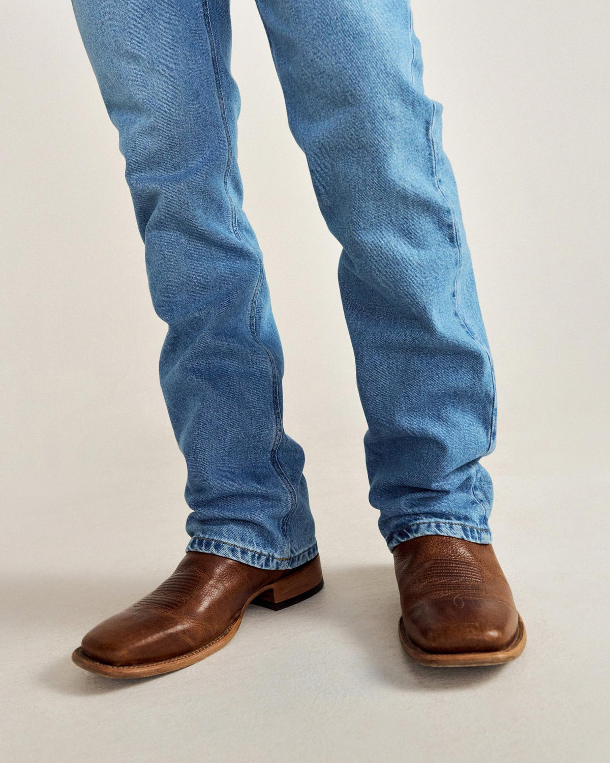 Person wearing light blue jeans and brown leather cowboy boots standing on a light-colored surface.