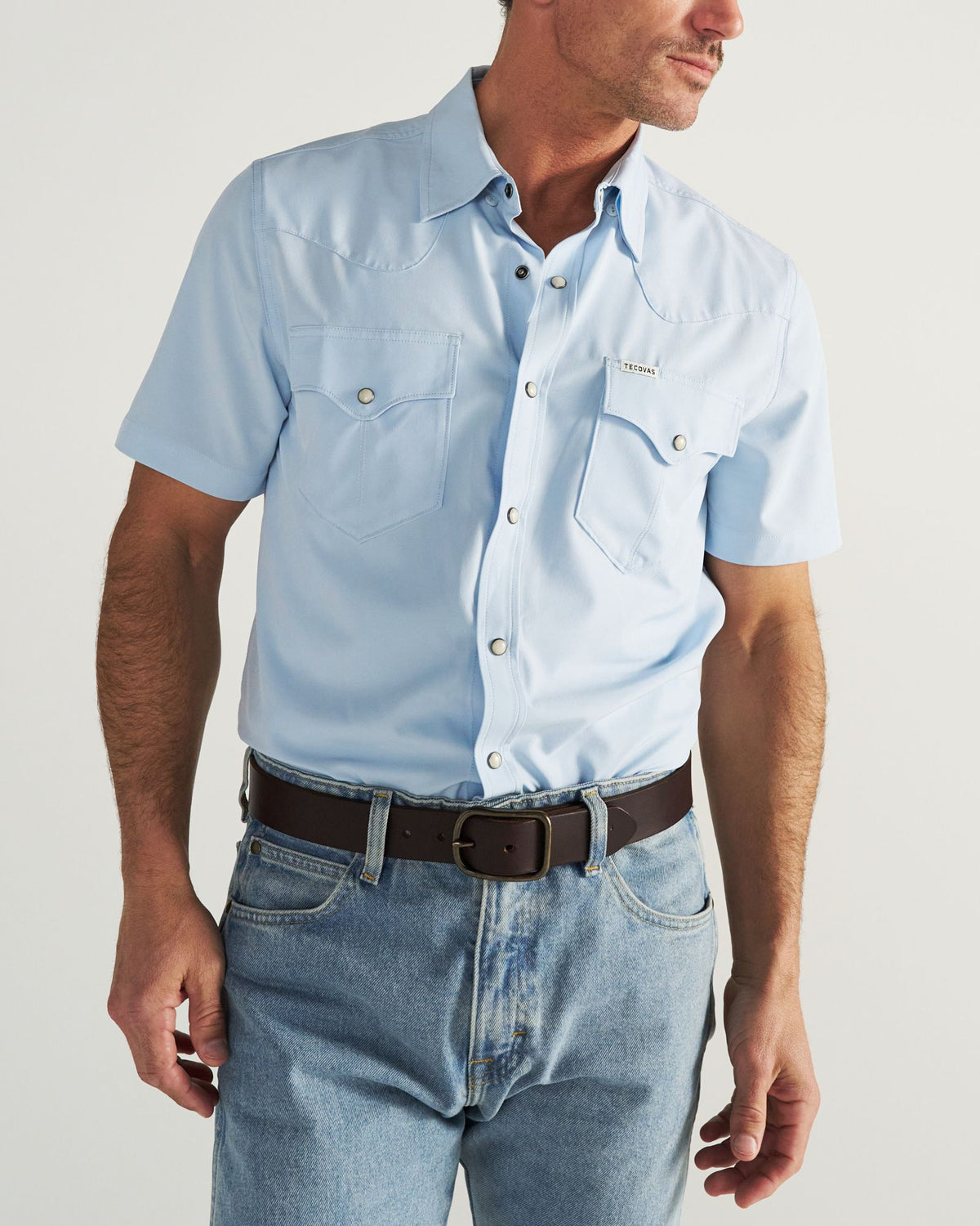 A man wearing a light blue short-sleeve button-up shirt with front pockets, blue jeans, and a black belt, standing against a plain background.