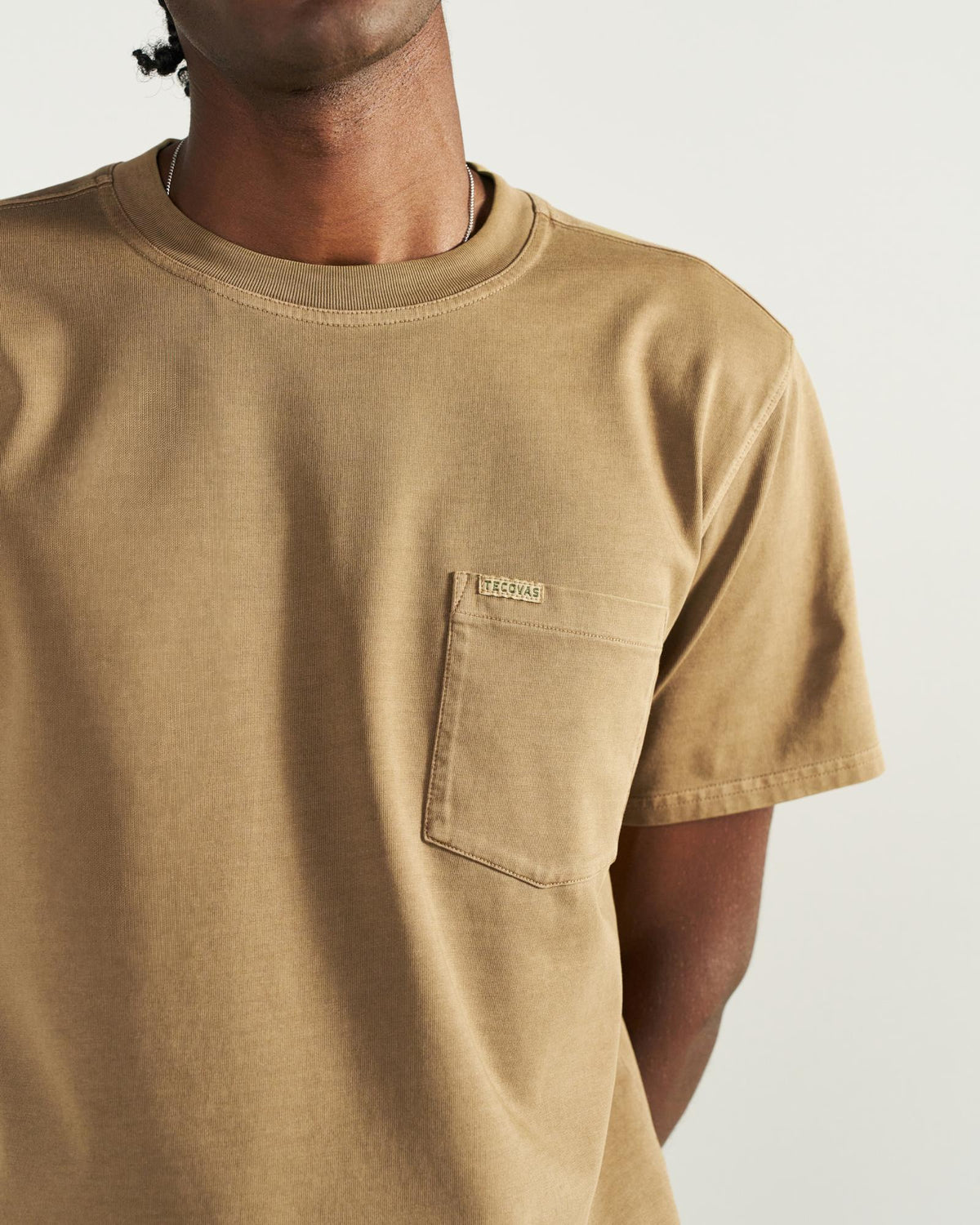 A person wearing a tan short-sleeve T-shirt with a chest pocket and small label, photographed from the shoulders to the waist against a plain background.