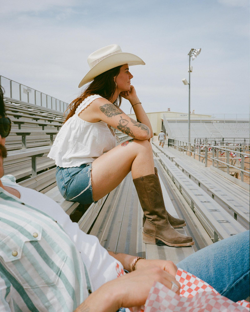 A woman in a cowboy hat, white sleeveless top, denim shorts, and boots sits on bleachers at an outdoor stadium; nearby, The Charlie Goat cafe’s logo peeks from the food held by someone in a striped shirt.