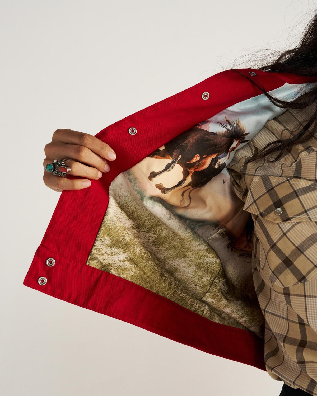 A person opens a red jacket to reveal an inner lining featuring an image of a running horse; the person wears a plaid shirt and a turquoise ring.