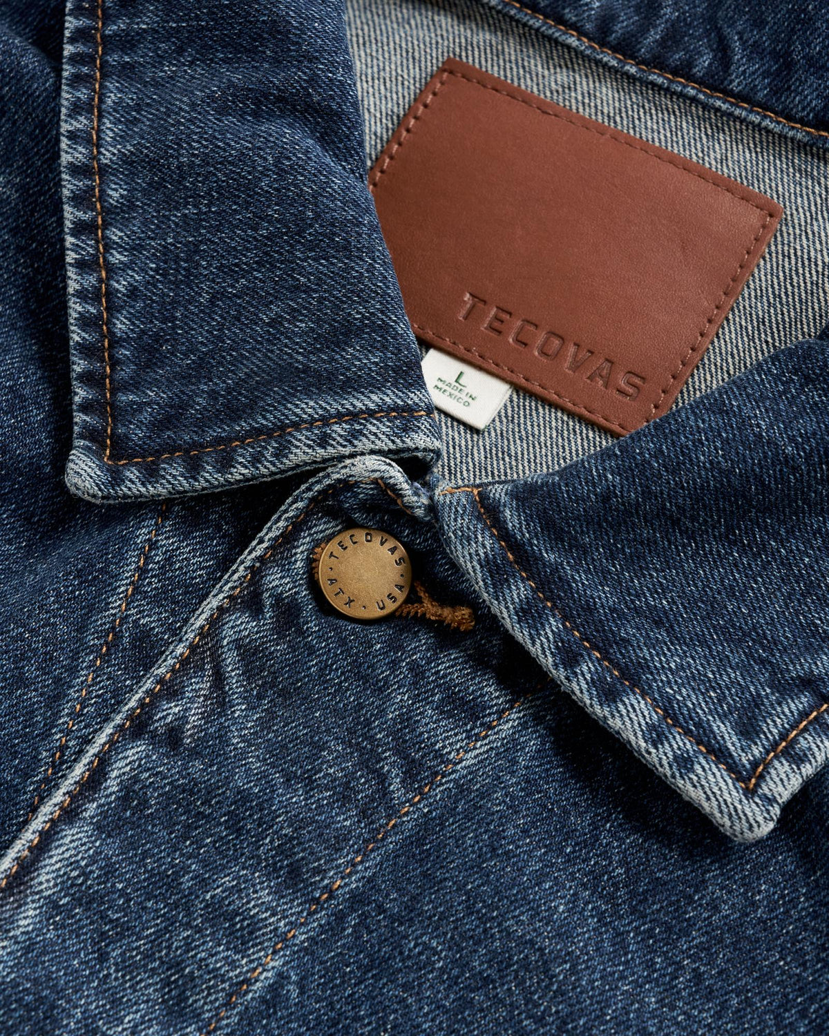Close-up of a blue denim jacket collar featuring a brown leather Tecovas brand patch, a size Large tag made in Mexico, and a brass button stamped with “Tecovas ATX USA.”.