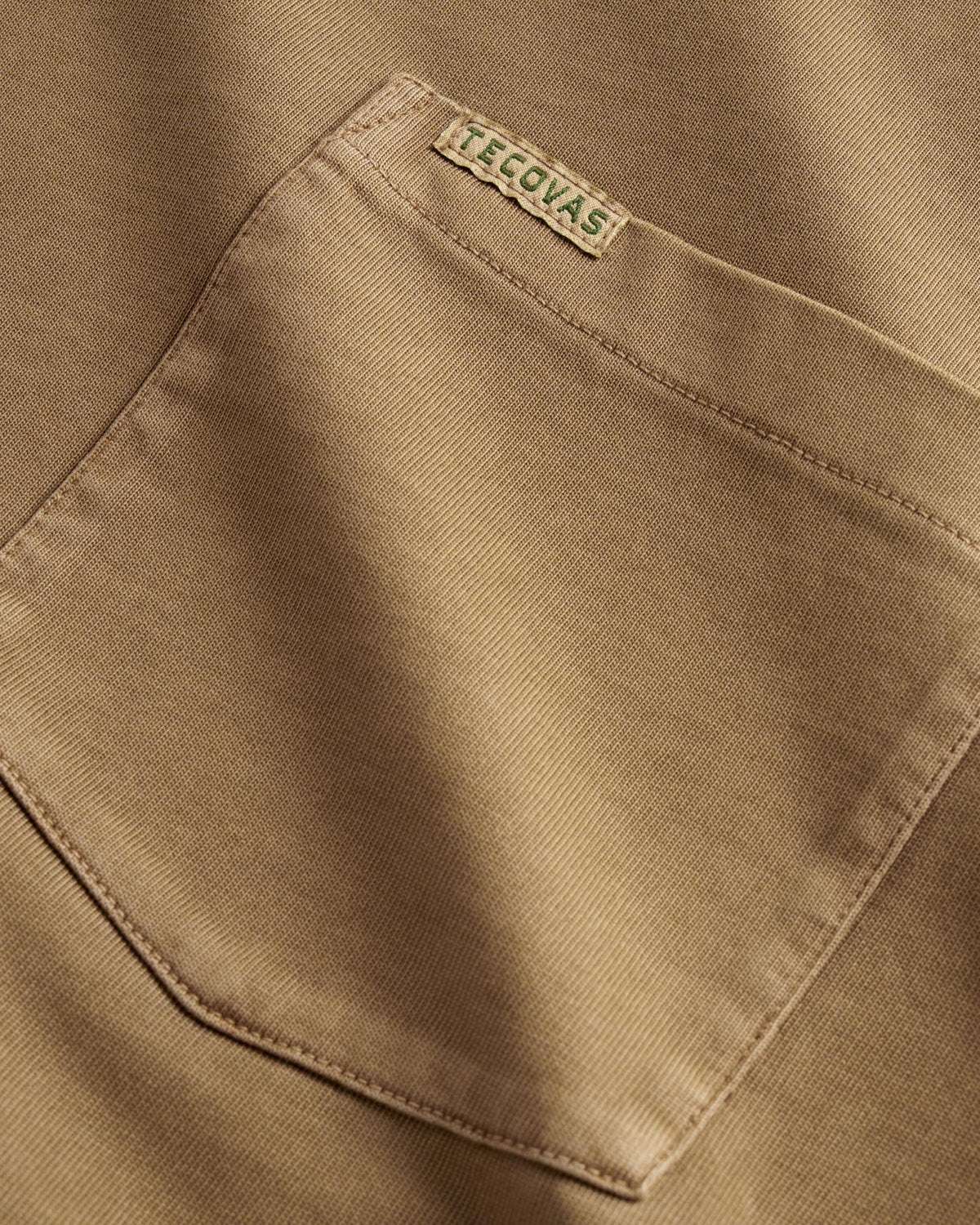 Close-up of a tan fabric shirt pocket with visible stitching, featuring a small rectangular label that reads “TECOVAS” sewn near the top edge.