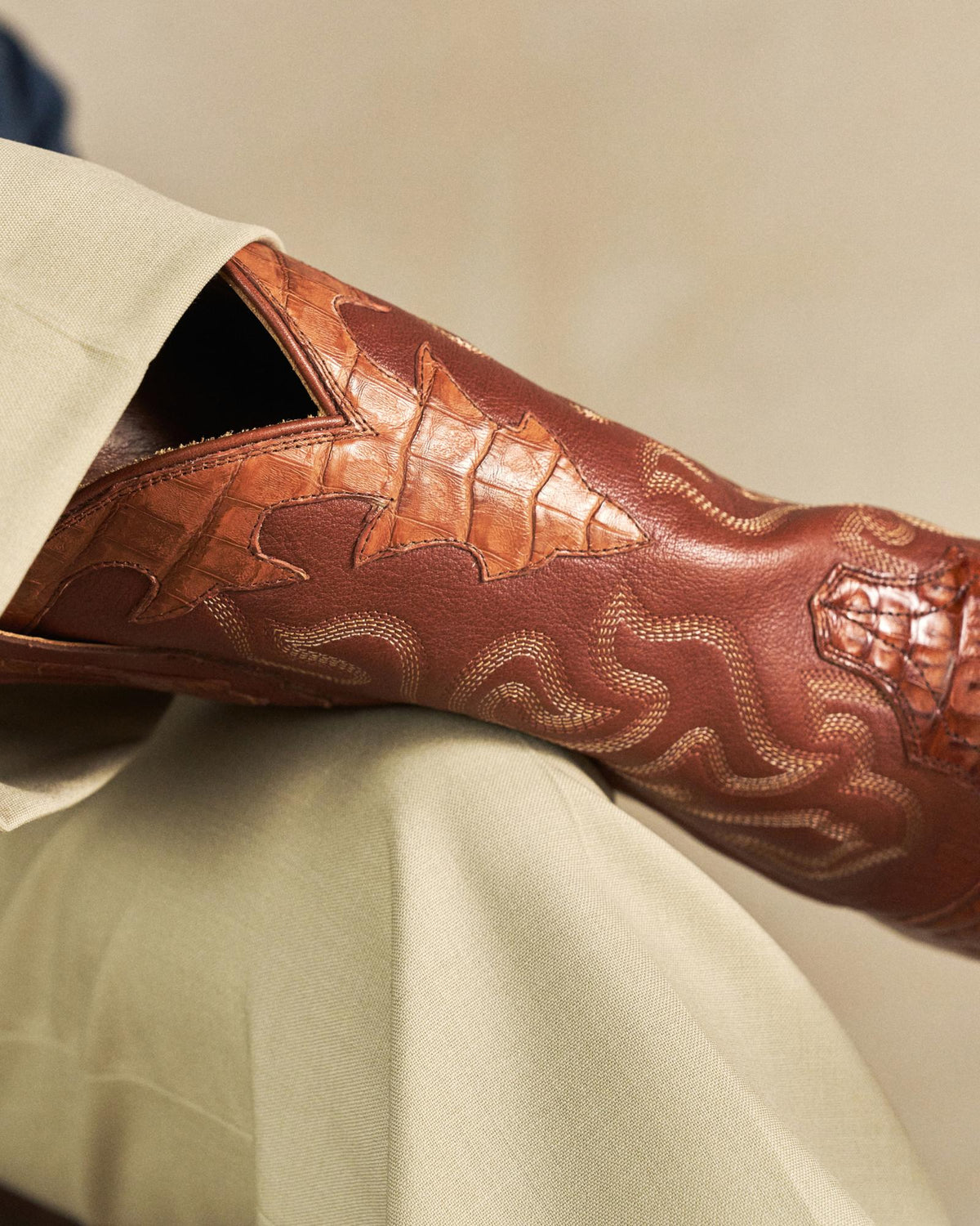 The Garrett in Pecan Hornback Caiman. Close-up of a brown leather Hornback Caiman cowboy boot with decorative stitching, resting on a crossed knee in light-colored pants.