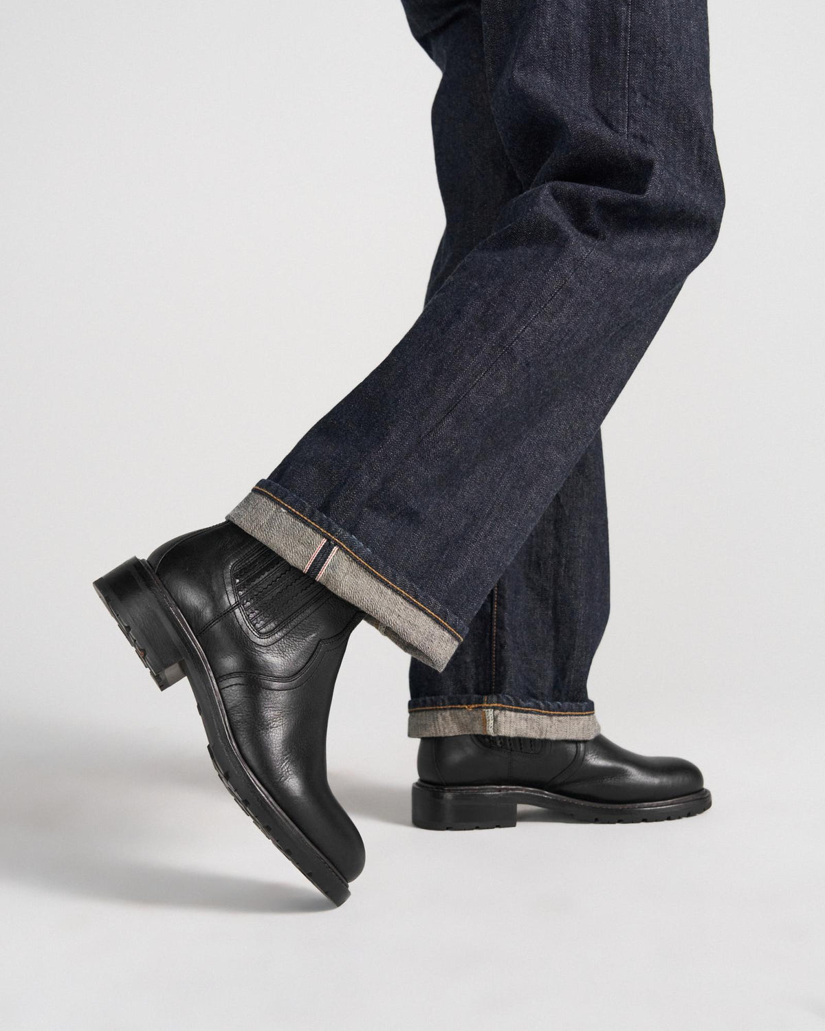 The Chelsea in Midnight Cowhide. Person wearing dark cuffed jeans and black leather boots, standing with one foot slightly raised against a plain background—the Midnight Cowhide boots add a refined edge to this timeless look.