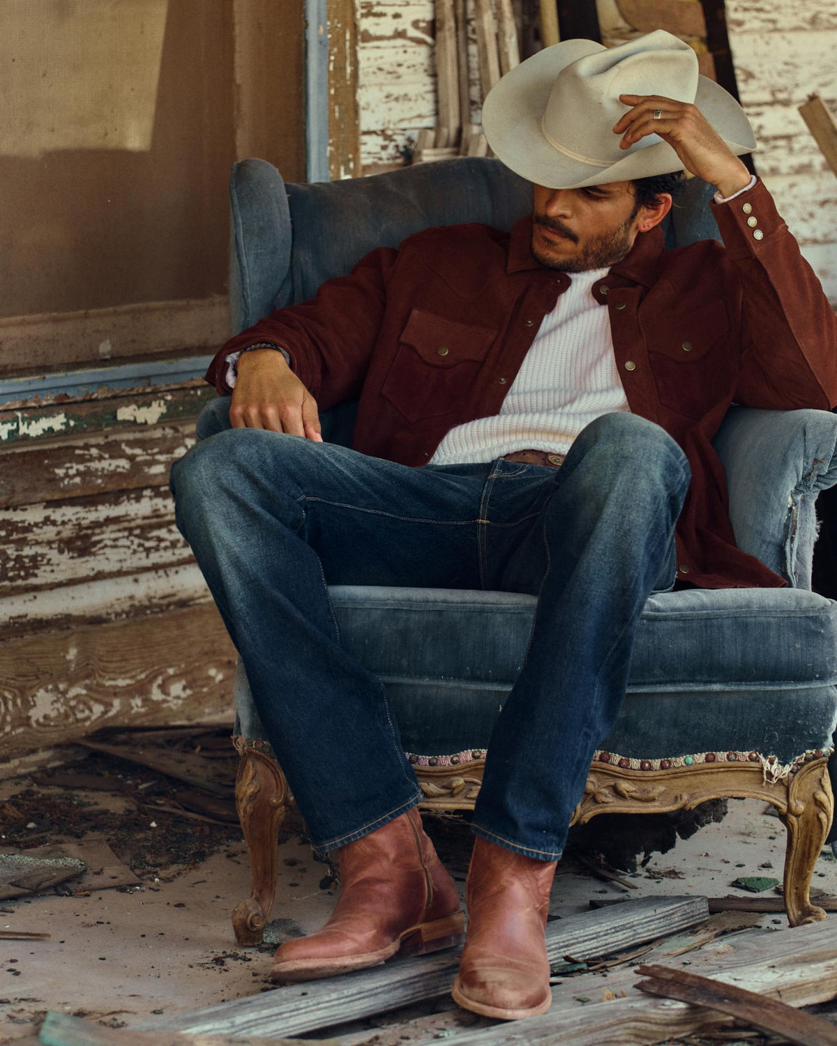 A man in a cowboy hat, brown jacket, jeans, and boots sits relaxed in a worn armchair outside a weathered building, savoring a glass of Scotch as he watches over The Dean Goat nearby.