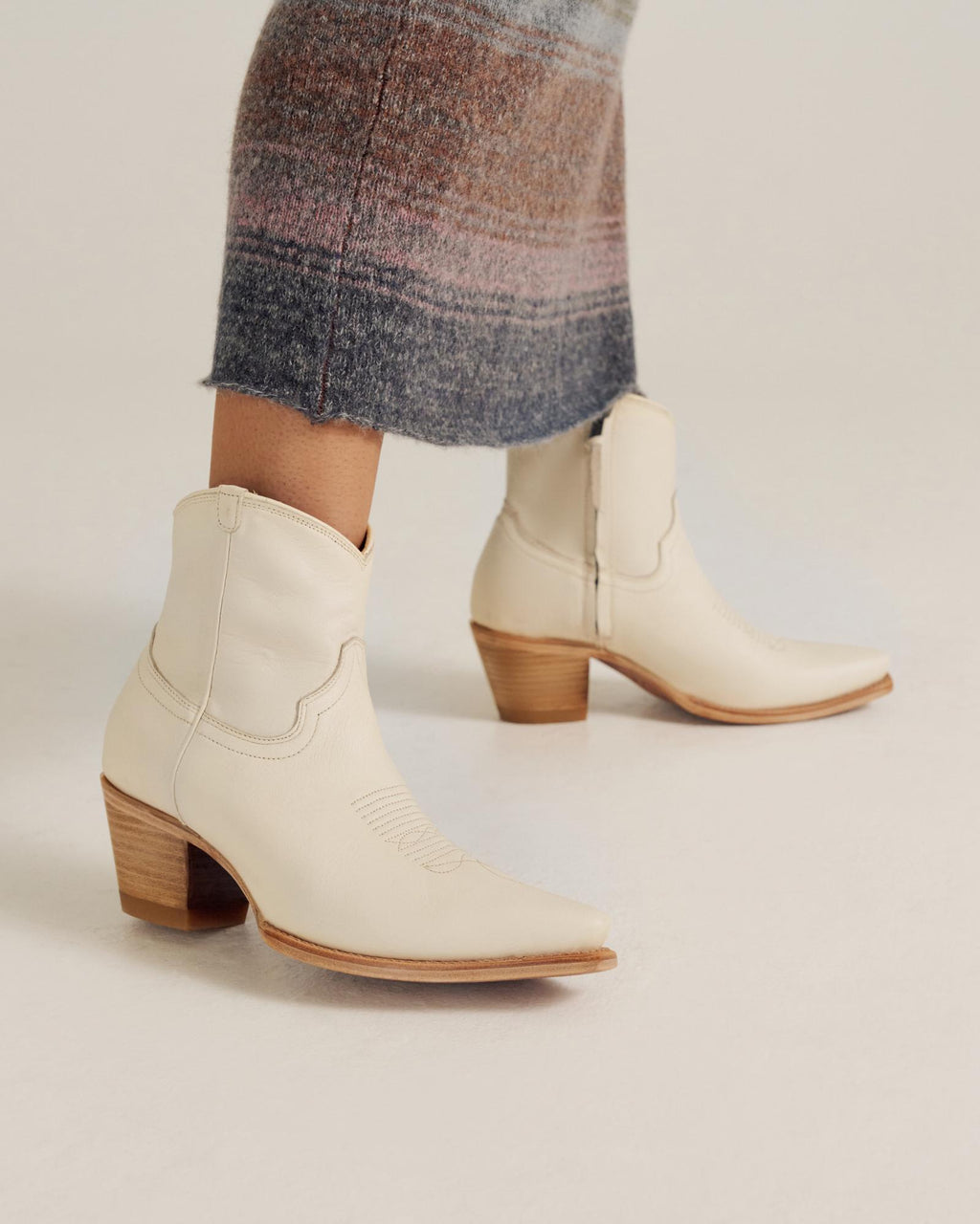 A person wearing a long, striped skirt and off-white, mid-calf western boots with side zippers and wooden heels.
