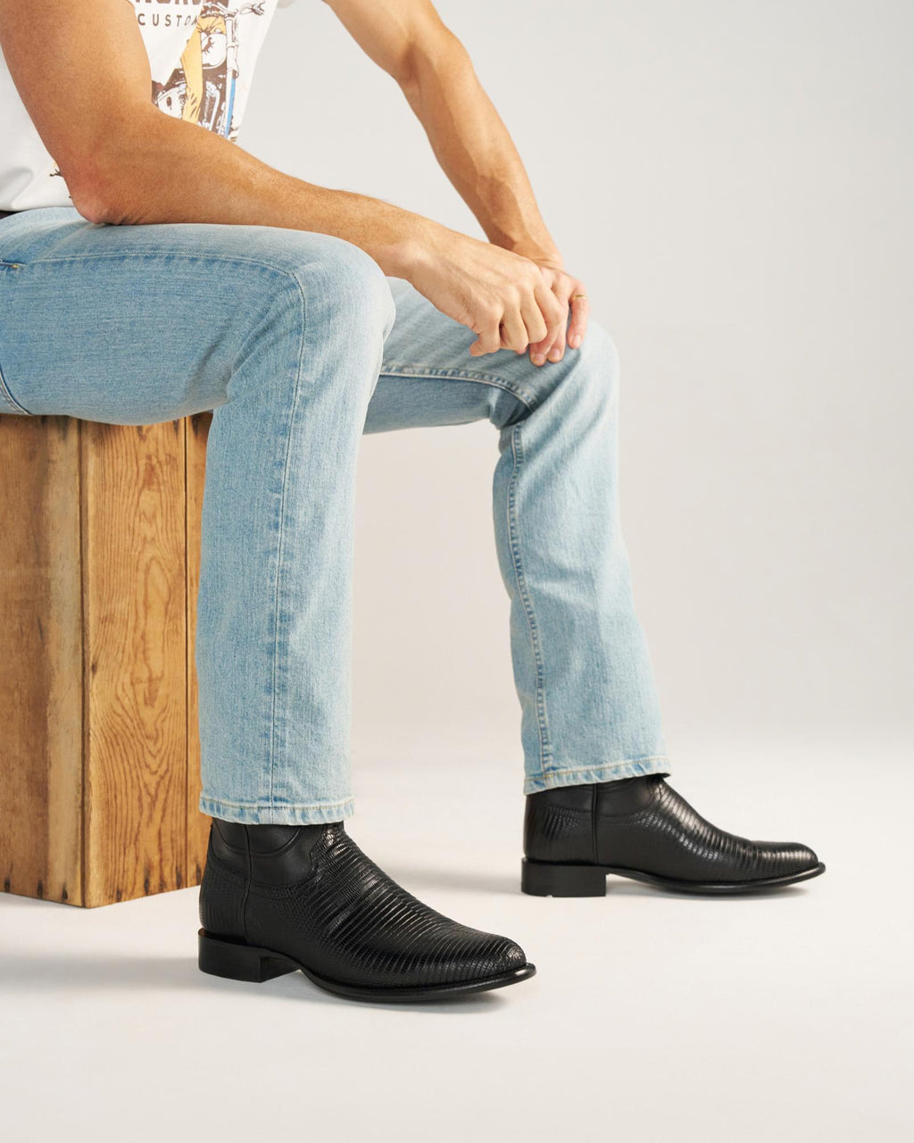 The Earl in Obsidian Lizard. A person wearing light blue jeans, a white T-shirt, and Obsidian black textured boots sits on a wooden box against a plain background.