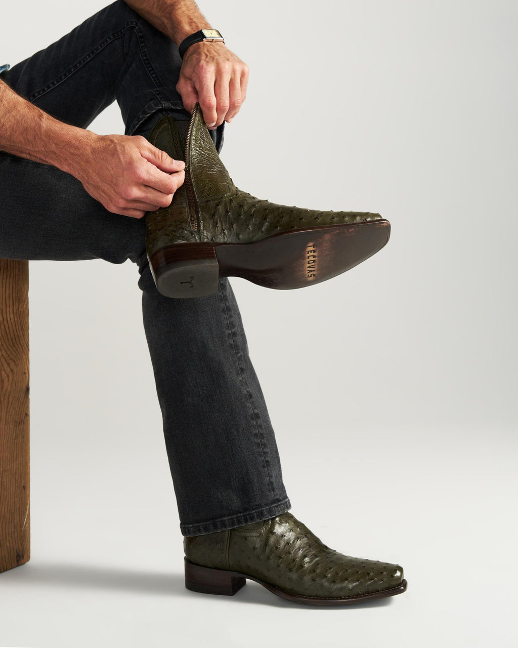 The Jack in Dark Forest Ostrich. A person in dark jeans zips up an olive green textured cowboy boot crafted from luxurious Dark Forest Ostrich leather while sitting on a wooden bench.