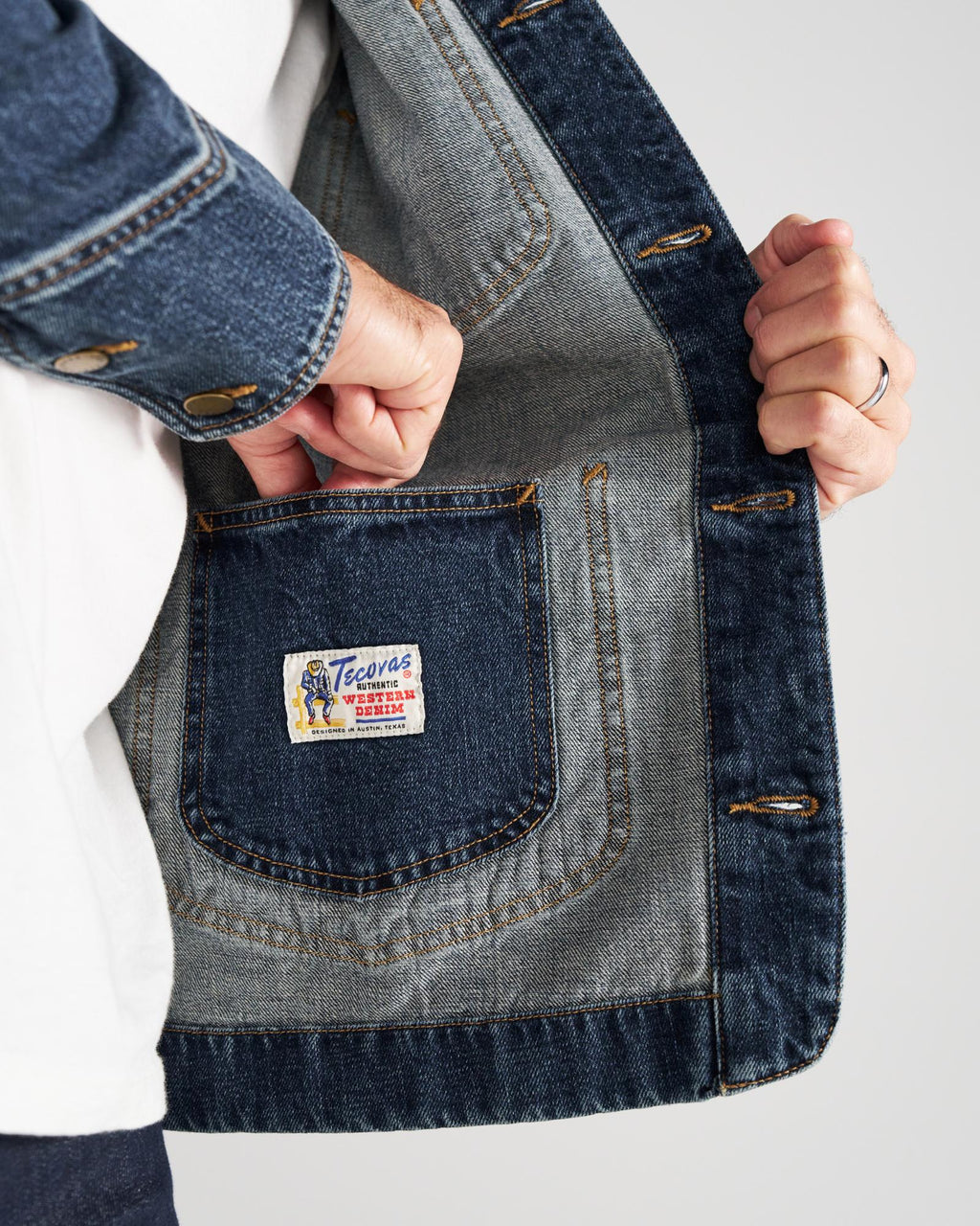 A person opens a denim jacket to show an inside pocket with a label that reads “Tecovas Western Goods.”.