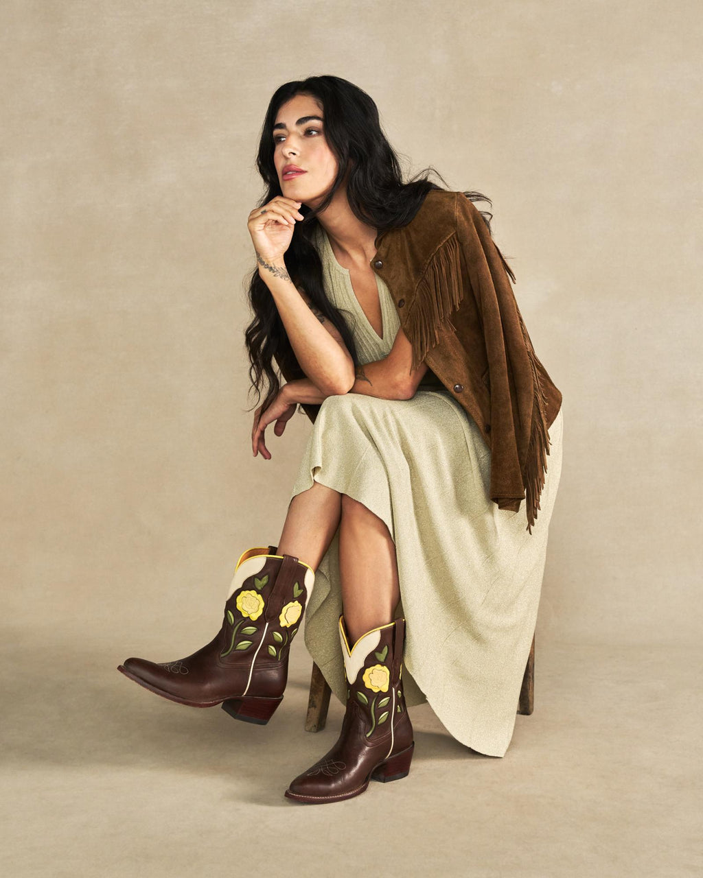 The Jolene in Truffle Cowhide. Woman seated on a chair, wearing a long beige dress, brown fringed jacket, and distinctive brown cowboy boots with yellow flower designs inspired by The Jolene Cowhide collection, against a plain beige background.