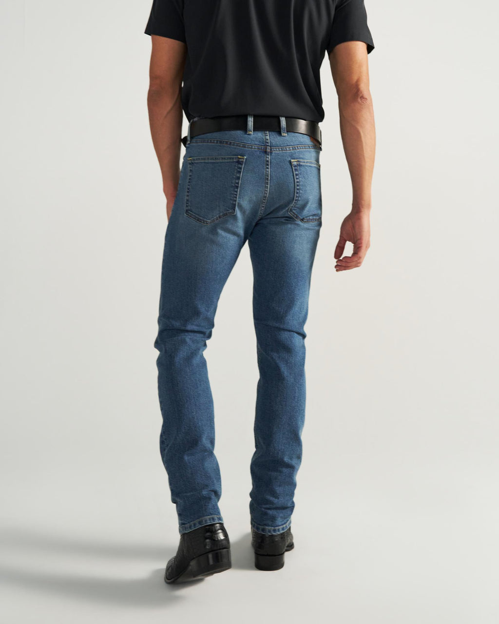 Man wearing a black shirt, blue jeans, black belt, and black shoes, standing with his back to the camera against a plain background.