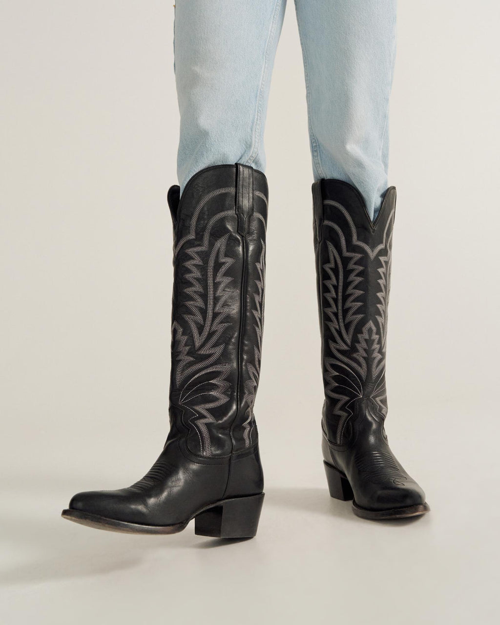 A person wearing light blue jeans and black western cowboy boots stands on a light-colored surface.