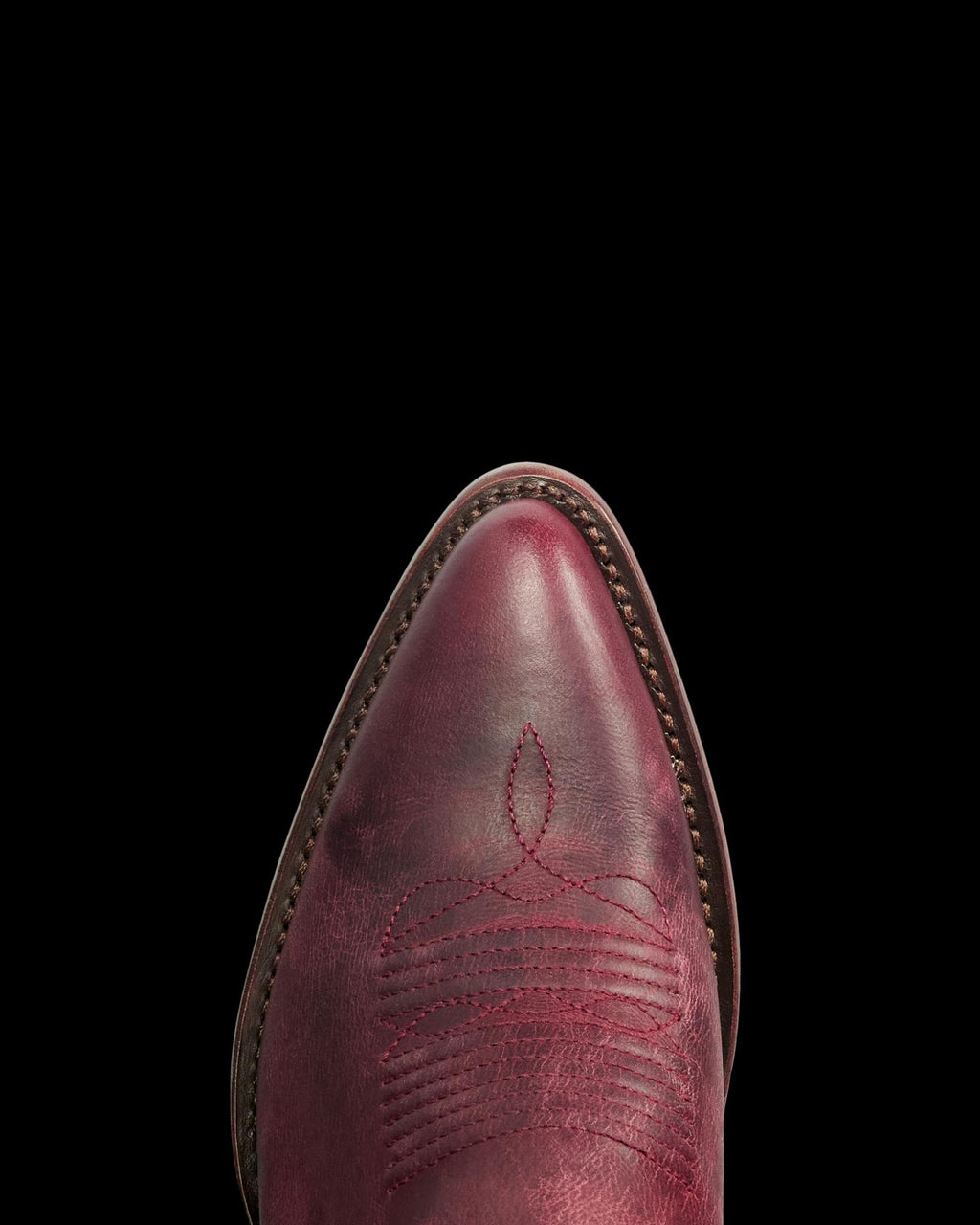 The Abby in Wine Goat. Close-up of the pointed toe of a red leather cowboy boot from The Abby Goat collection, featuring decorative stitching and a brown welt, shown against a white background.
