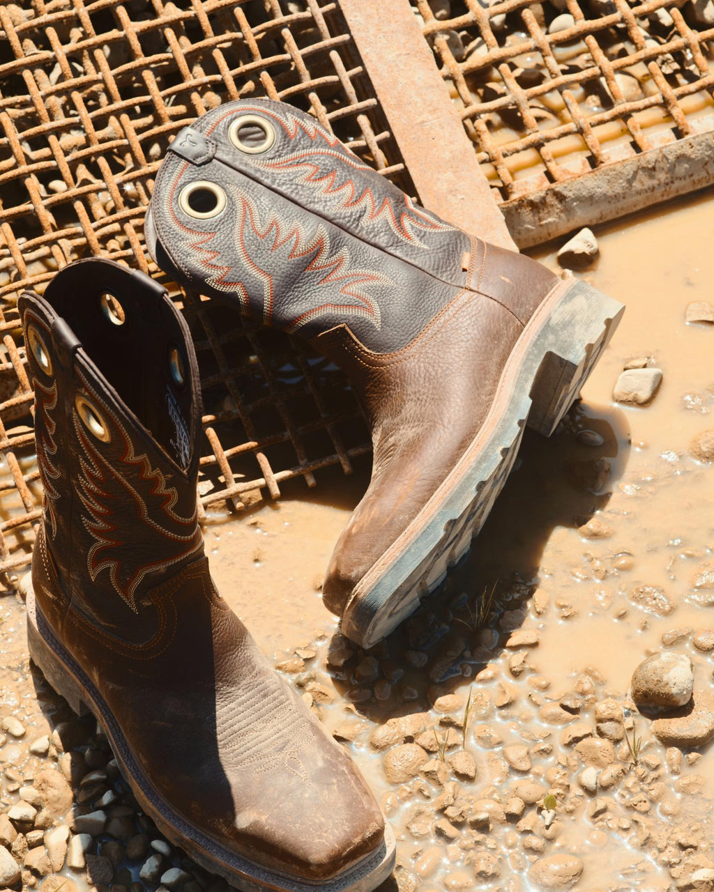 LongHaul Waterproof Square Composite Toe Work Boot in Tobacco / Black Cowhide. A pair of worn Tobacco / Black Cowhide cowboy boots with decorative stitching are placed on wet ground near a metal grate and muddy puddle.