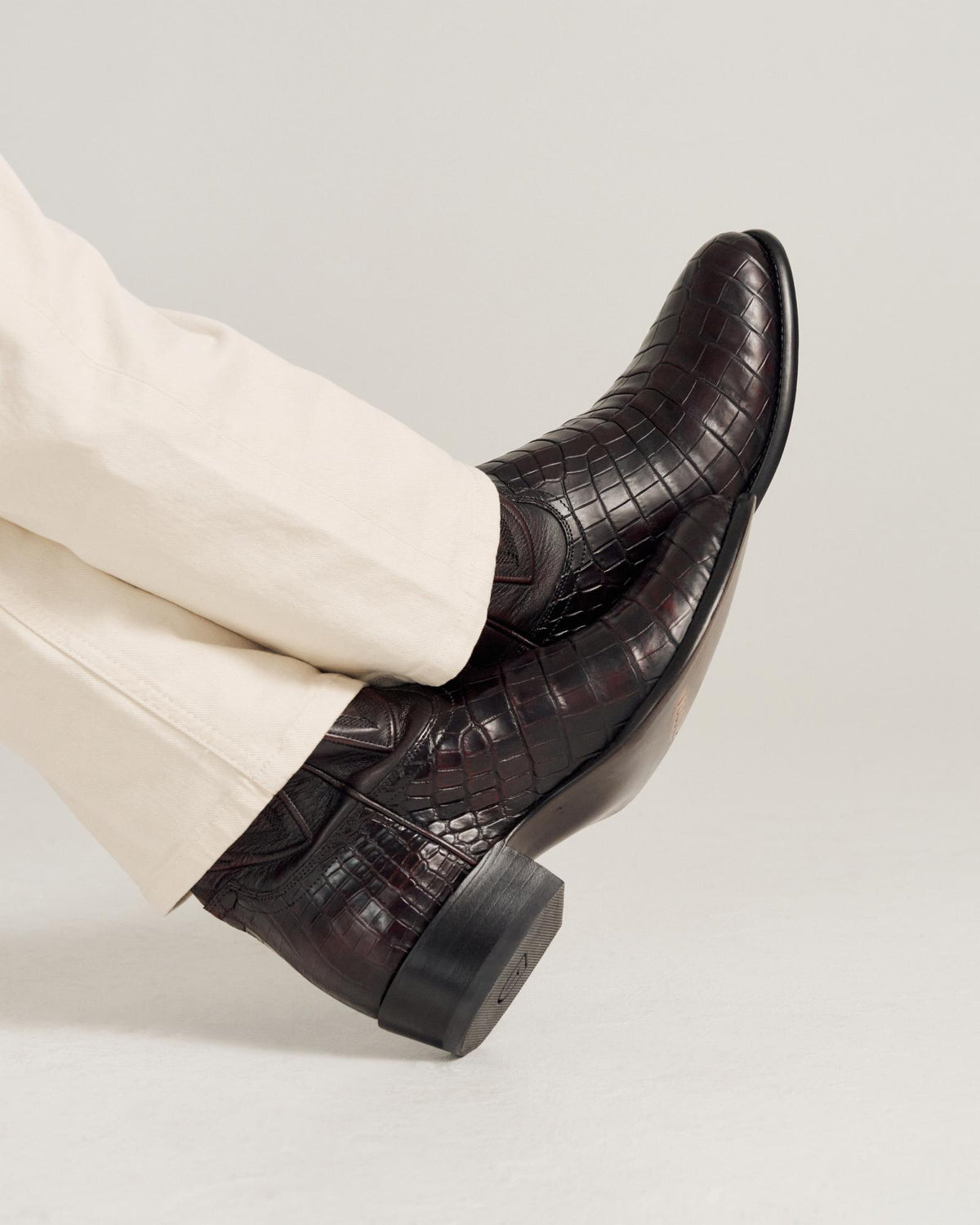 A person wearing light-colored pants and dark brown, crocodile-pattern leather boots with a low heel, resting their legs crossed.