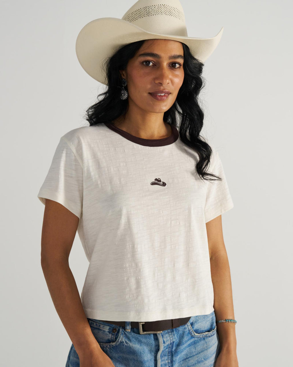Woman in a white cowboy hat and white t-shirt with a small emblem, wearing blue jeans, stands against a neutral background.