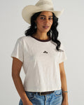 Woman in a white cowboy hat and white t-shirt with a small emblem, wearing blue jeans, stands against a neutral background.