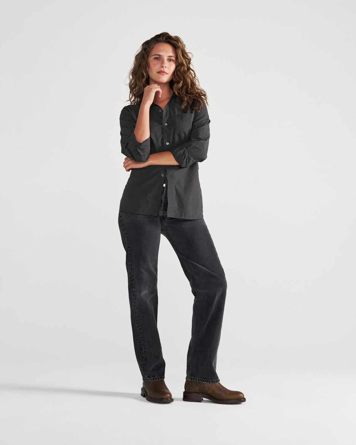 A person with long curly hair, wearing a dark button-up shirt, dark jeans, and brown boots, stands with one hand on their chin against a plain white background—evoking the relaxed yet stylish atmosphere of The Chelsea Cowhide cafe.