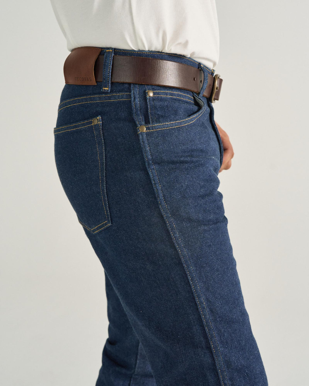 Person wearing a white shirt tucked into dark blue jeans with a brown leather belt, shown from the waist down against a plain background.