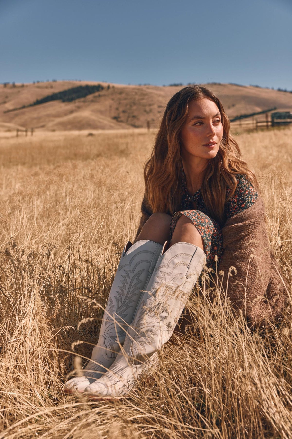 Woman sitting in a grassy field against a backdrop of hills, wearing a floral dress, brown shawl, and white boots.