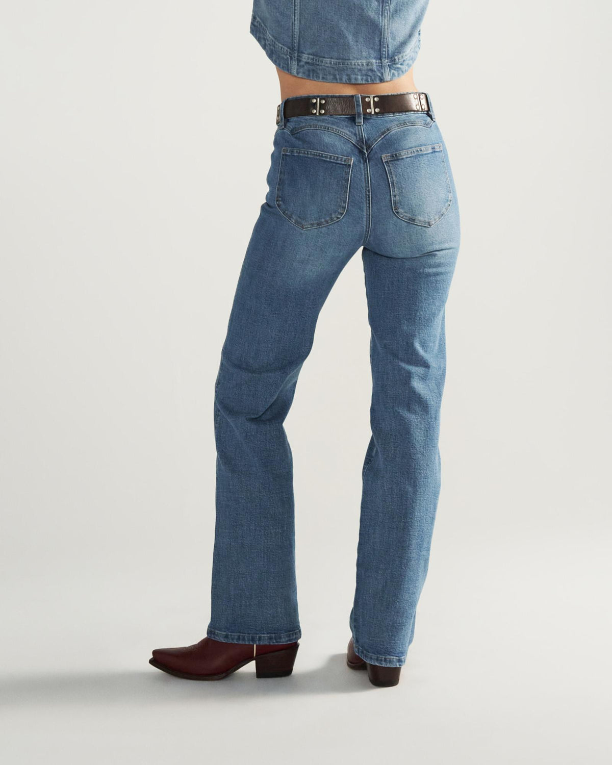 Person wearing blue jeans, a black belt, and brown heeled boots, photographed from behind against a plain background.