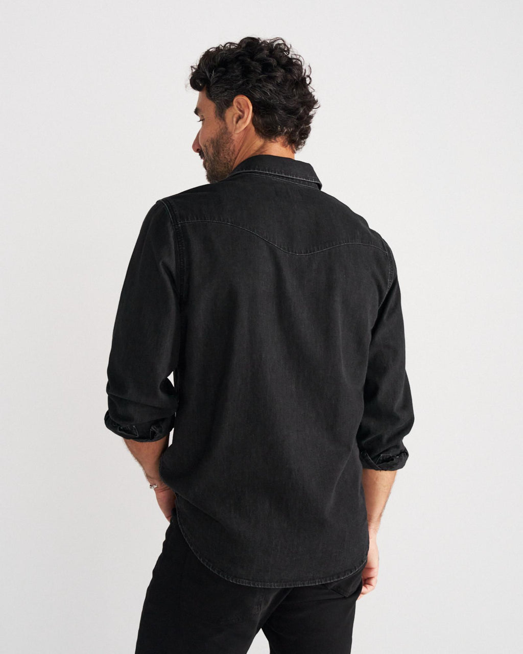 A man with dark curly hair is viewed from behind, wearing a black long-sleeved button-down shirt and black pants, standing against a plain white background.