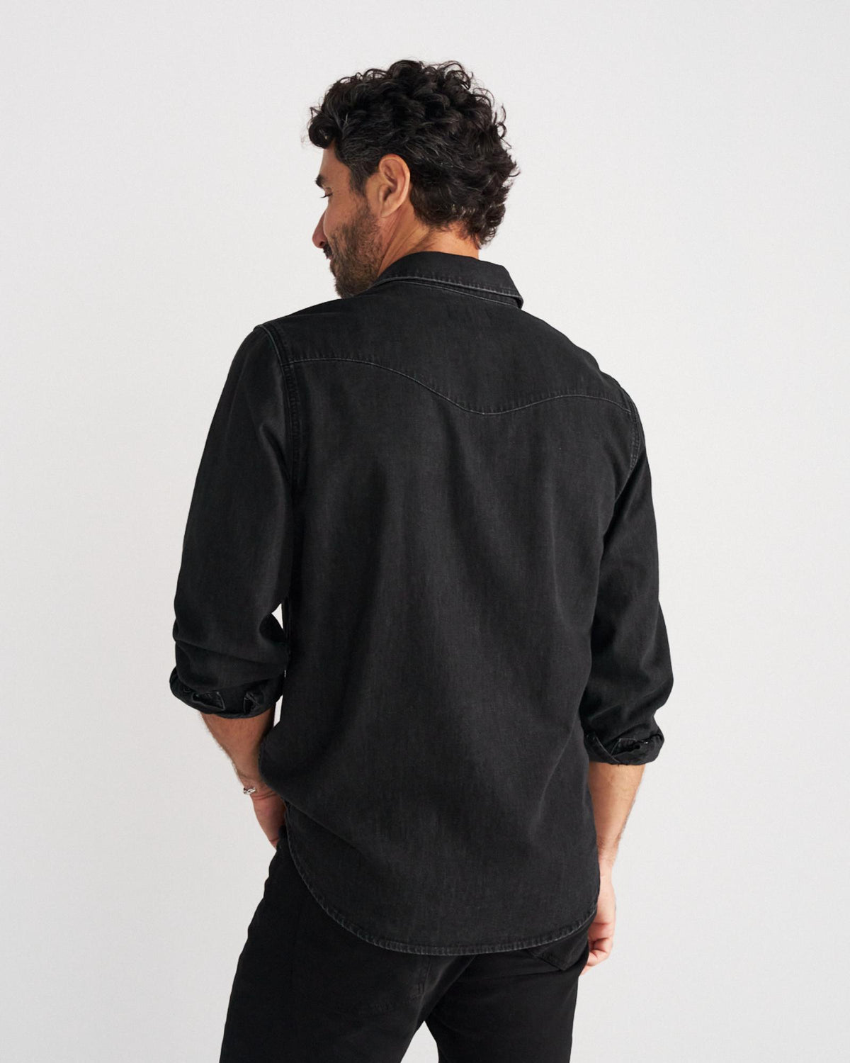 A man with dark curly hair is viewed from behind, wearing a black long-sleeved button-down shirt and black pants, standing against a plain white background.