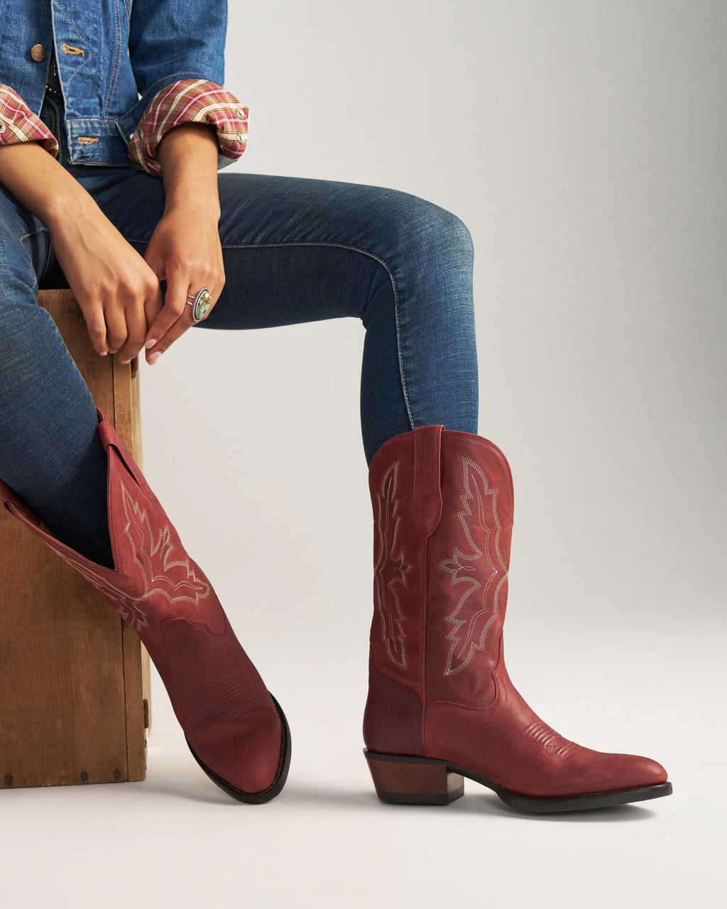 The Britt in Redwood Goat. A person wearing a denim jacket, jeans, and striking red cowboy boots sits on a wooden box from The Britt Goat, hands resting on their knee.