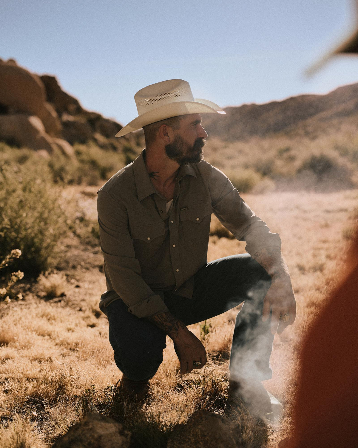 A man wearing a cowboy hat and button-up shirt kneels on dry grass in a desert landscape, with rocks and hills in the background.