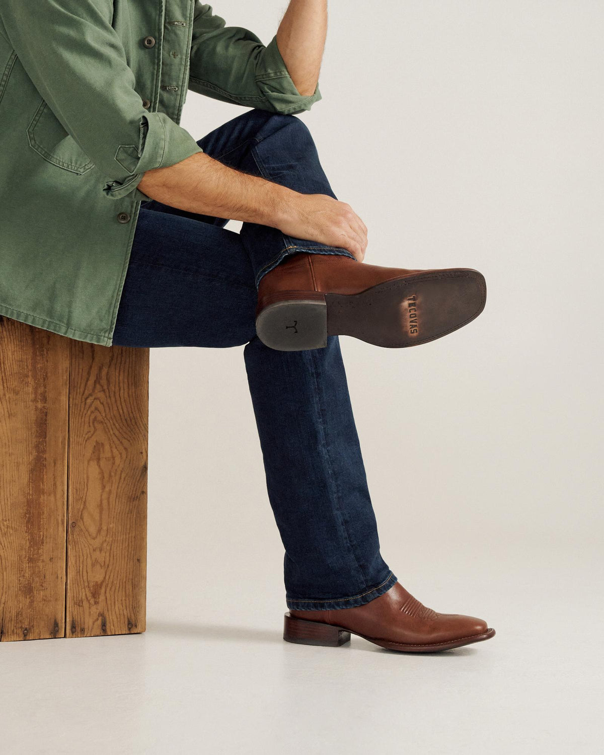 A person in a green jacket and blue jeans sits on a wooden block with legs crossed, wearing brown cowboy boots.