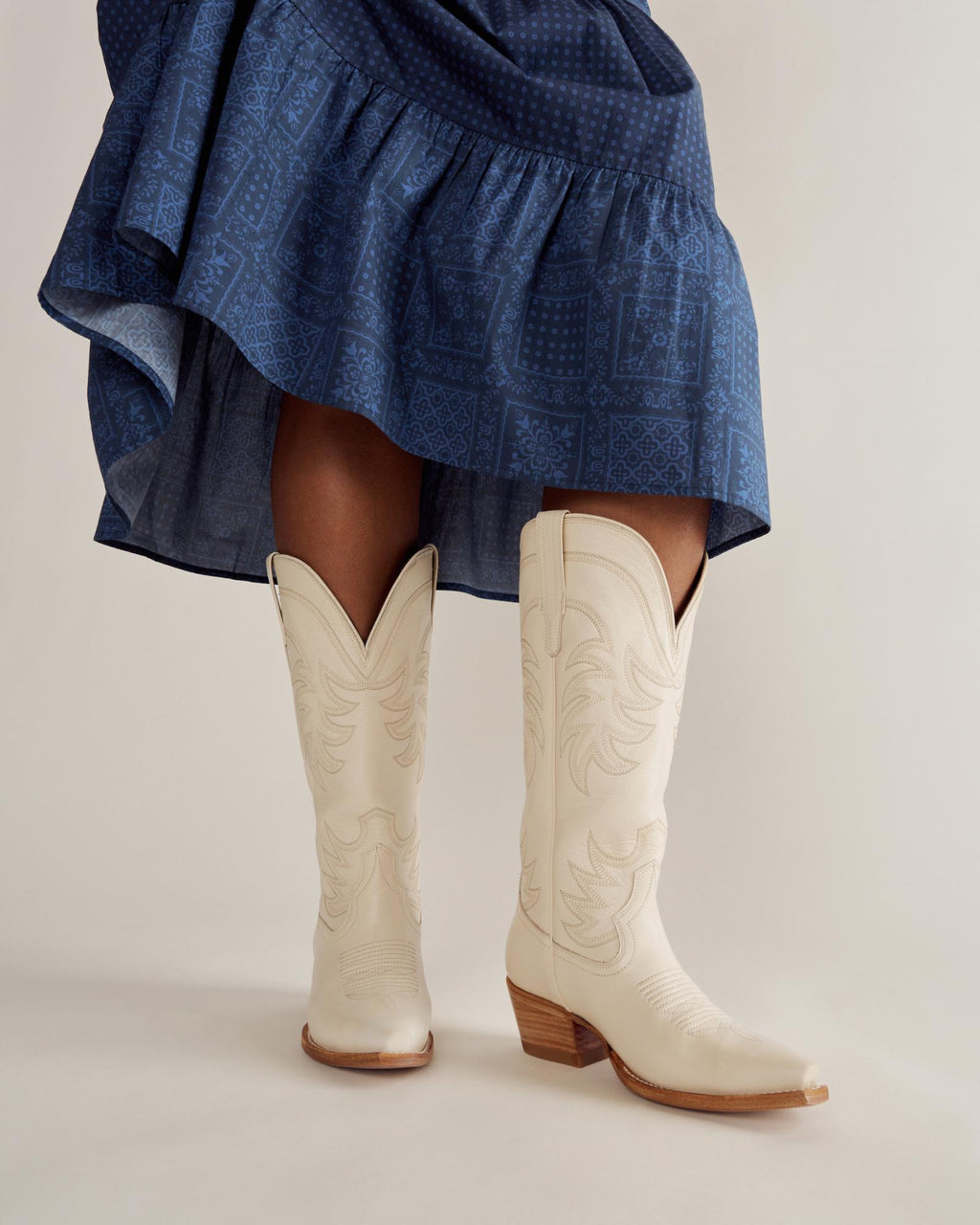 A person wearing a blue patterned skirt and white cowboy boots with a wooden heel, standing on a light-colored surface.
