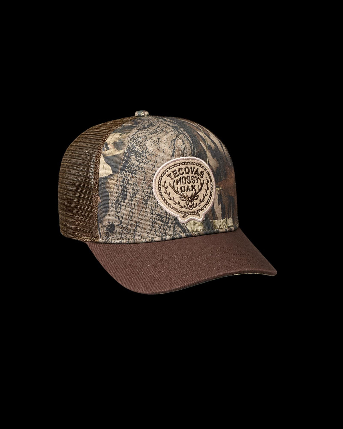 A brown and camouflage trucker hat with a mesh back and a round patch reading “Tecovas Mossy Oak” on the front.