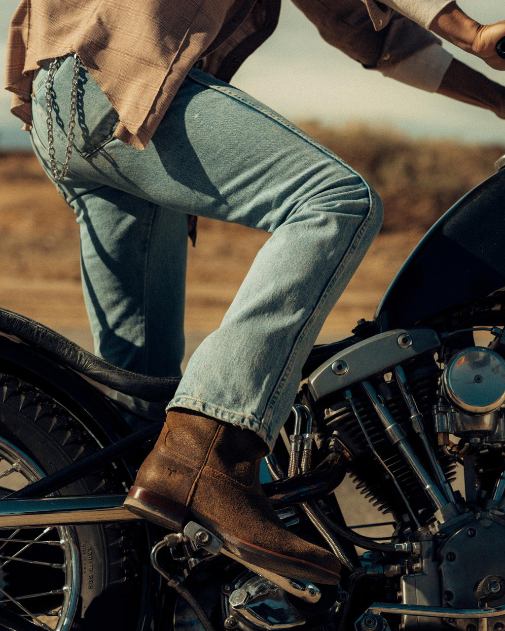 The Glen in Dustwood Roughout. Person wearing light blue jeans and brown boots sits on a vintage motorcycle, The Glen Roughout boots perfectly complementing the rugged desert landscape in the background.