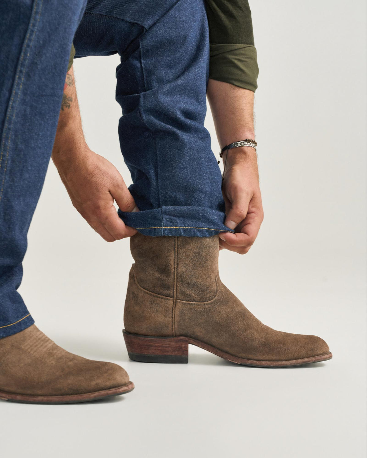 The Johnny in Gravel Suede. A person in blue jeans and a green shirt cuffs their jean leg over a brown Gravel Suede boot with a wooden heel.