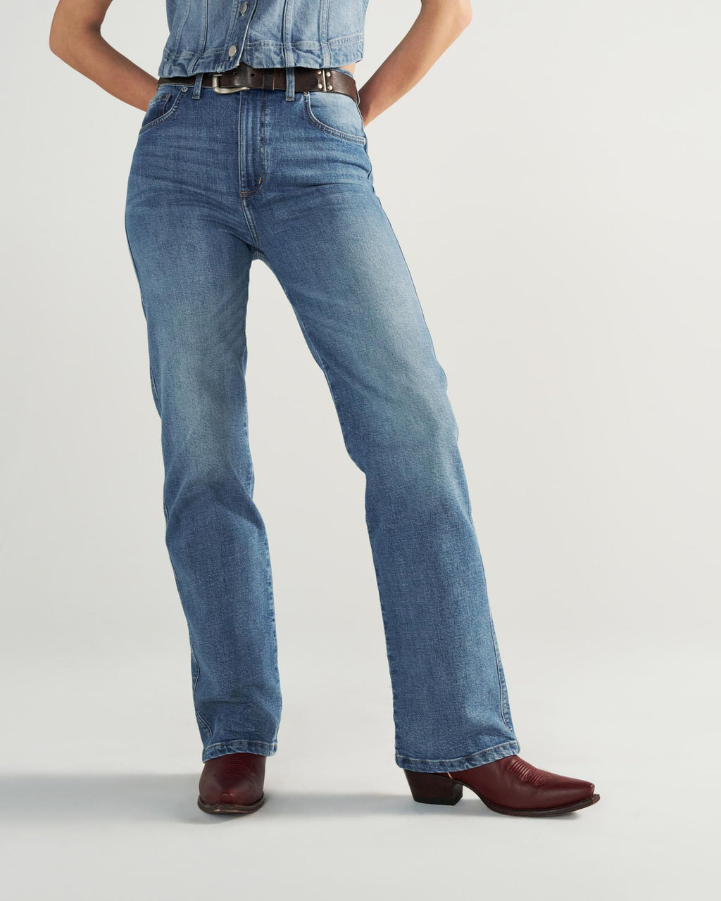 Person wearing blue jeans, a tucked-in denim shirt, a black belt, and burgundy cowboy boots, standing against a plain light background.