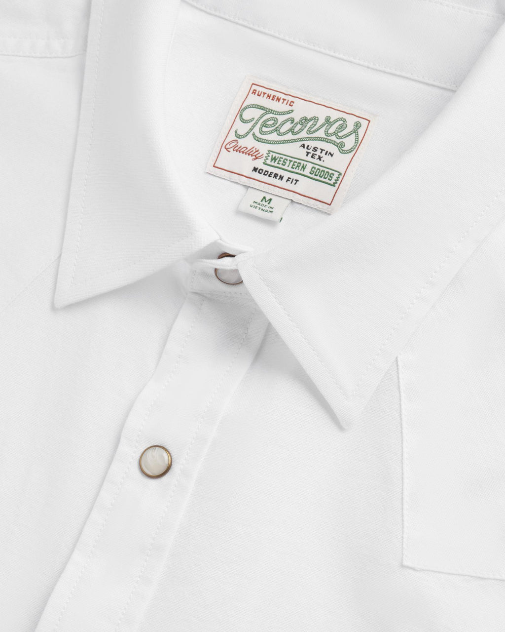 Close-up of a white collared shirt with a “Tecovas” brand label and pearl snap buttons, showing detail of stitching and fabric texture.