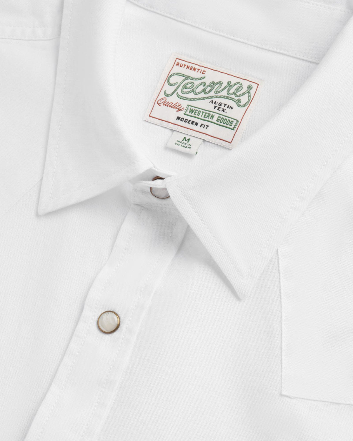 Close-up of a white collared shirt with a “Tecovas” brand label and pearl snap buttons, showing detail of stitching and fabric texture.