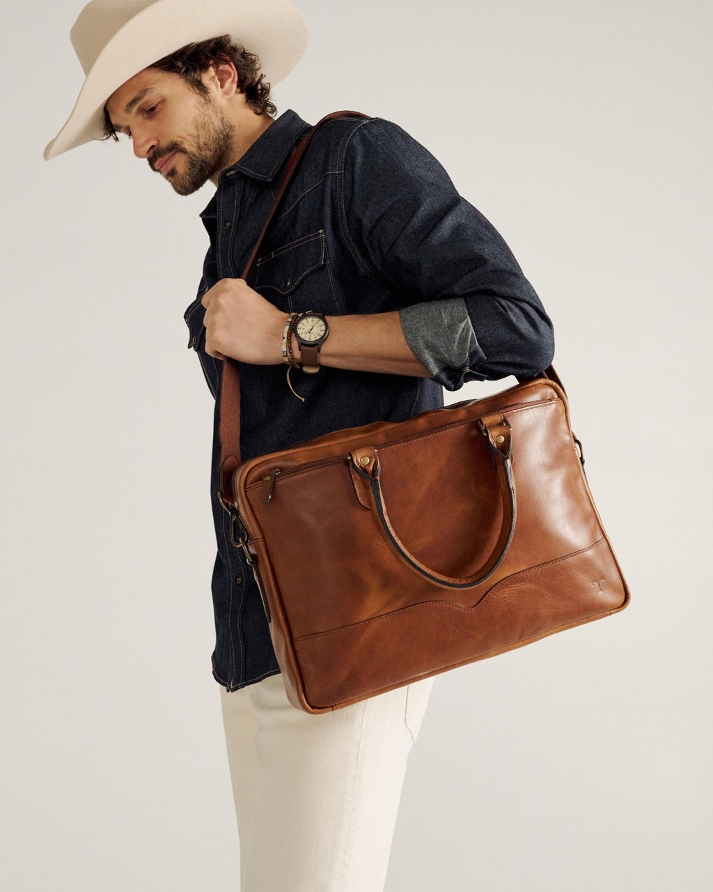 A man wearing a cowboy hat, dark denim shirt, and light pants carries a brown leather shoulder bag and wears a wristwatch.