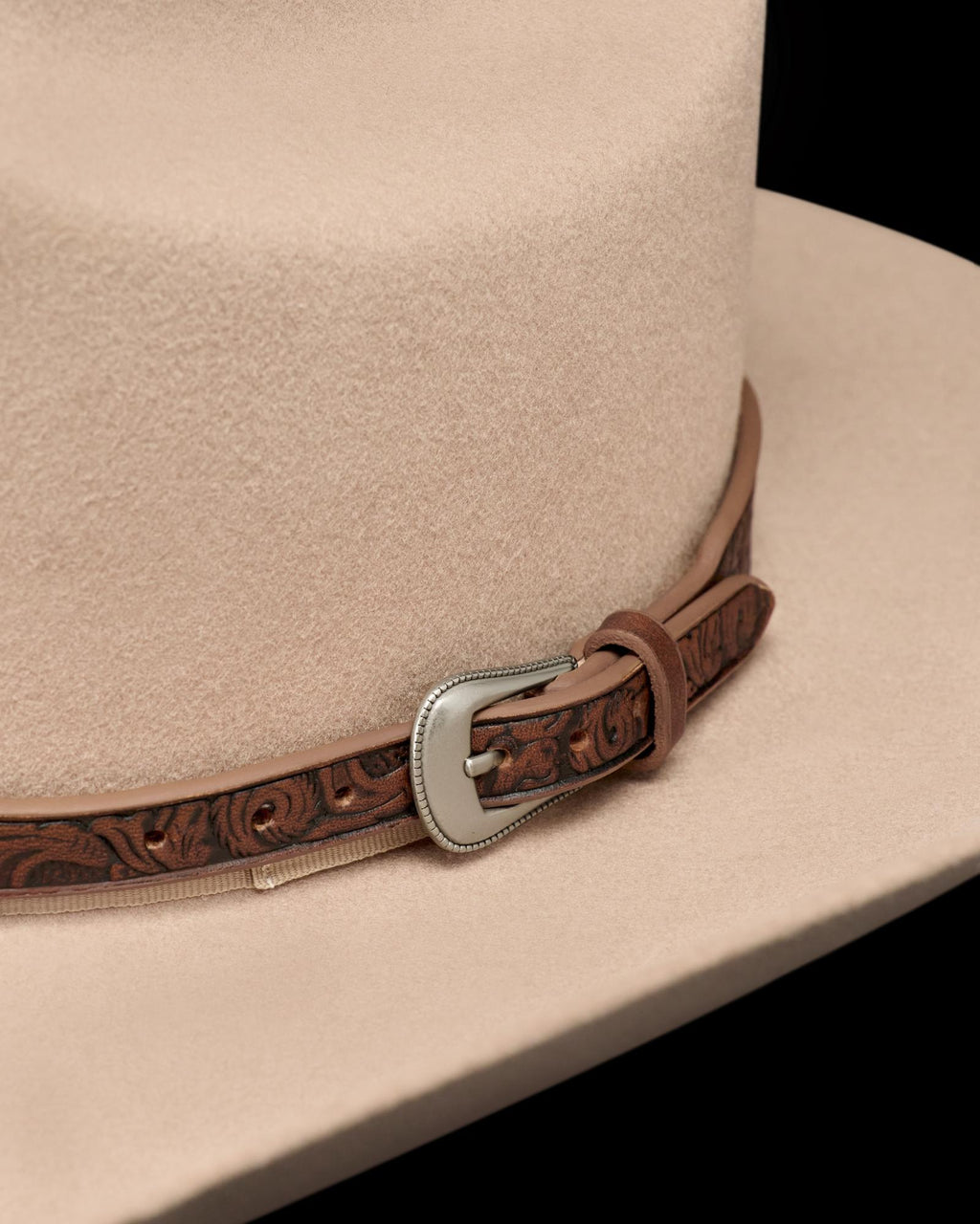 Close-up of a beige cowboy hat featuring a brown leather band with tooled patterns and a decorative silver buckle.