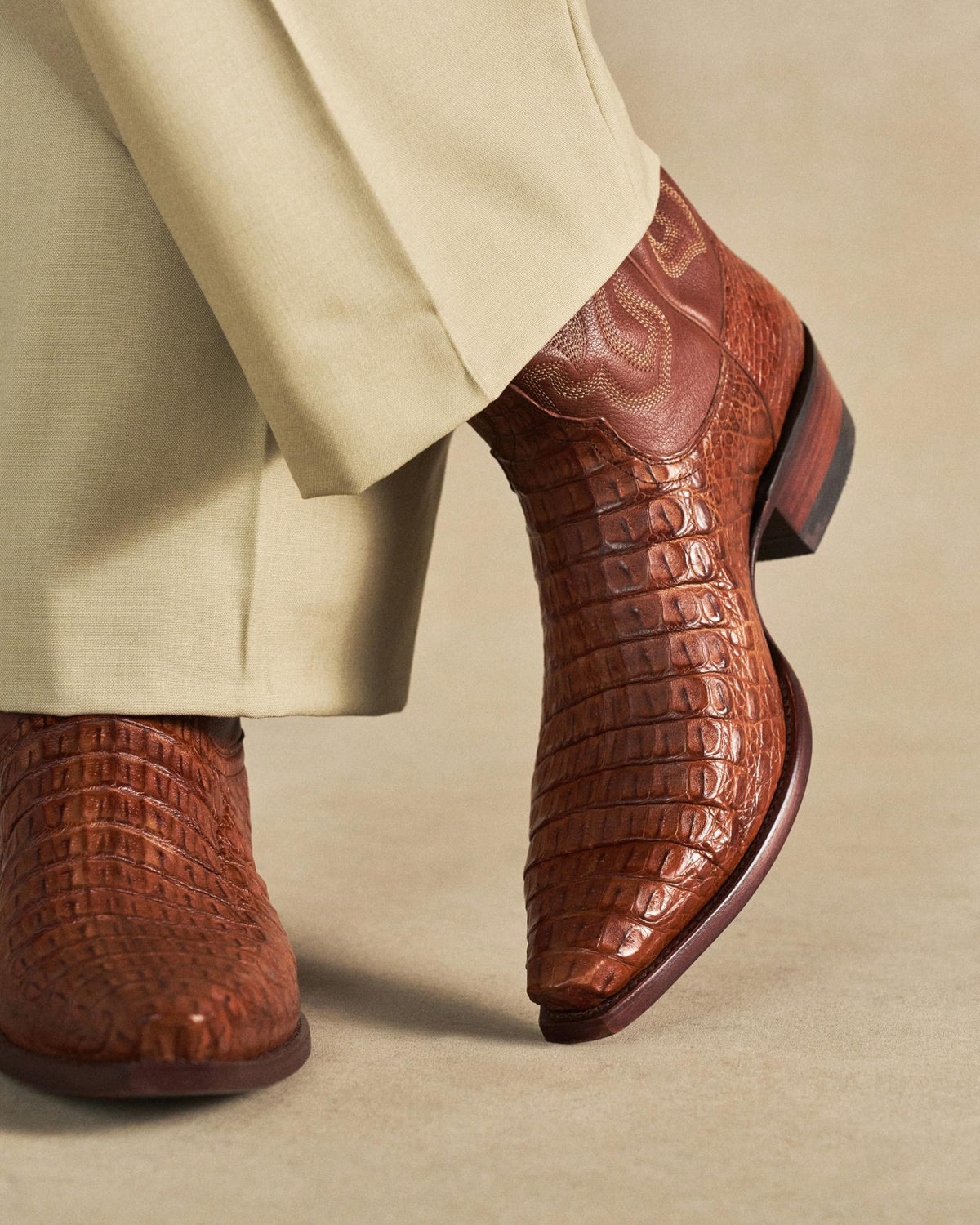 The Garrett in Pecan Hornback Caiman. A person wearing beige pants and brown textured cowboy boots, specifically The Garrett Hornback Caiman, stands on a light tan surface.