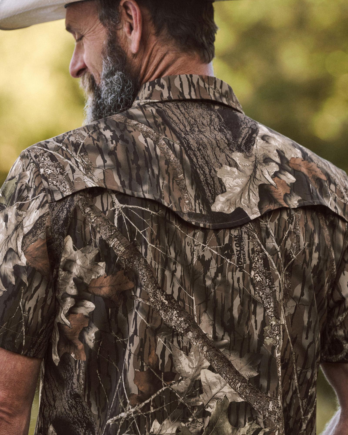 Man wearing a short-sleeved camouflage shirt with a leaf and branch pattern, standing outdoors and facing away from the camera.