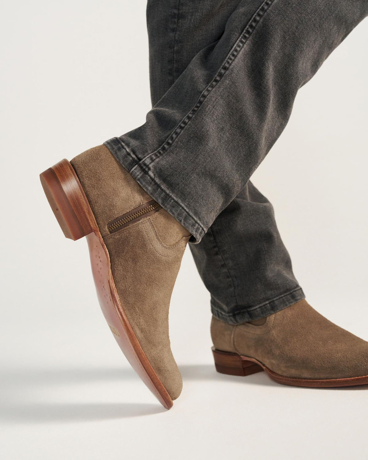 A person wearing gray jeans and The Dean Suede ankle boots in Granite, featuring light brown suede and wooden soles, stands against a plain background.
