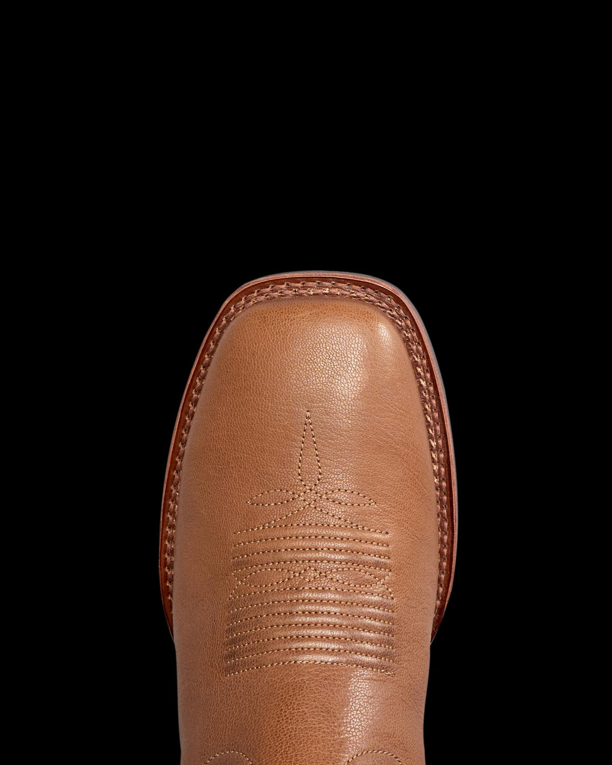 The Dusty in Mesa Goat. Close-up view of the toe of a tan leather cowboy boot from The Dusty Goat, featuring decorative stitching and a square toe design, shown on a white background.