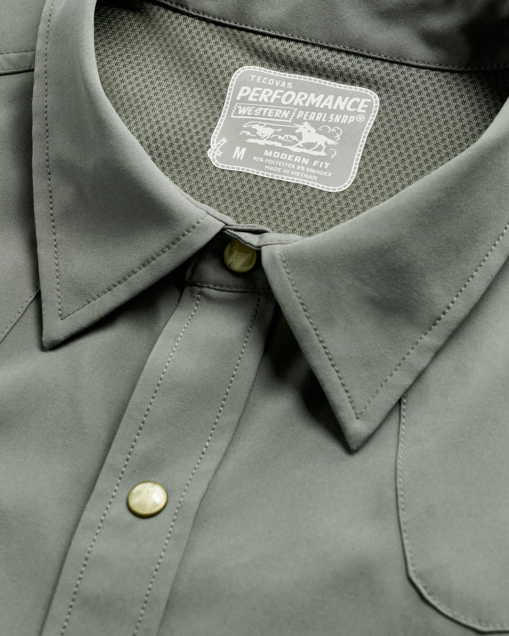 Close-up of a grey T-shirt featuring pearl snap buttons and a label reading "Tecovas Performance Western Pearl Snap, Medium, Modern Fit.