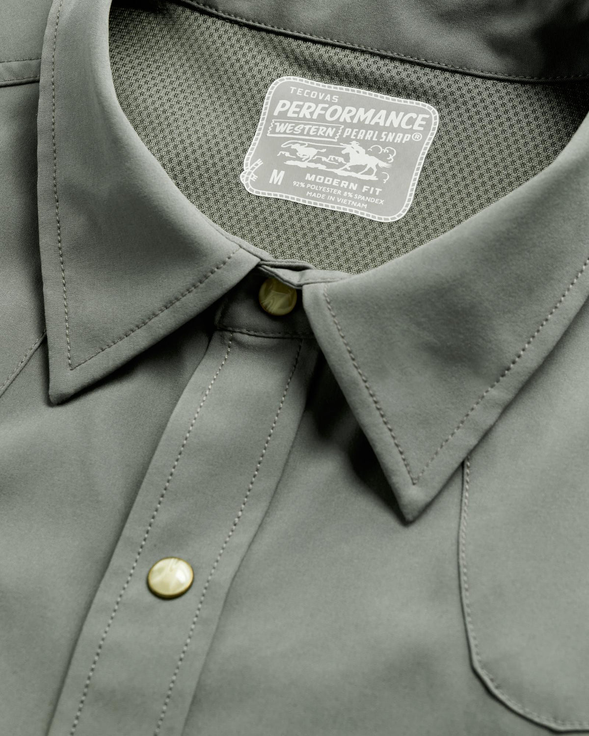 Close-up of a grey T-shirt featuring pearl snap buttons and a label reading "Tecovas Performance Western Pearl Snap, Medium, Modern Fit.