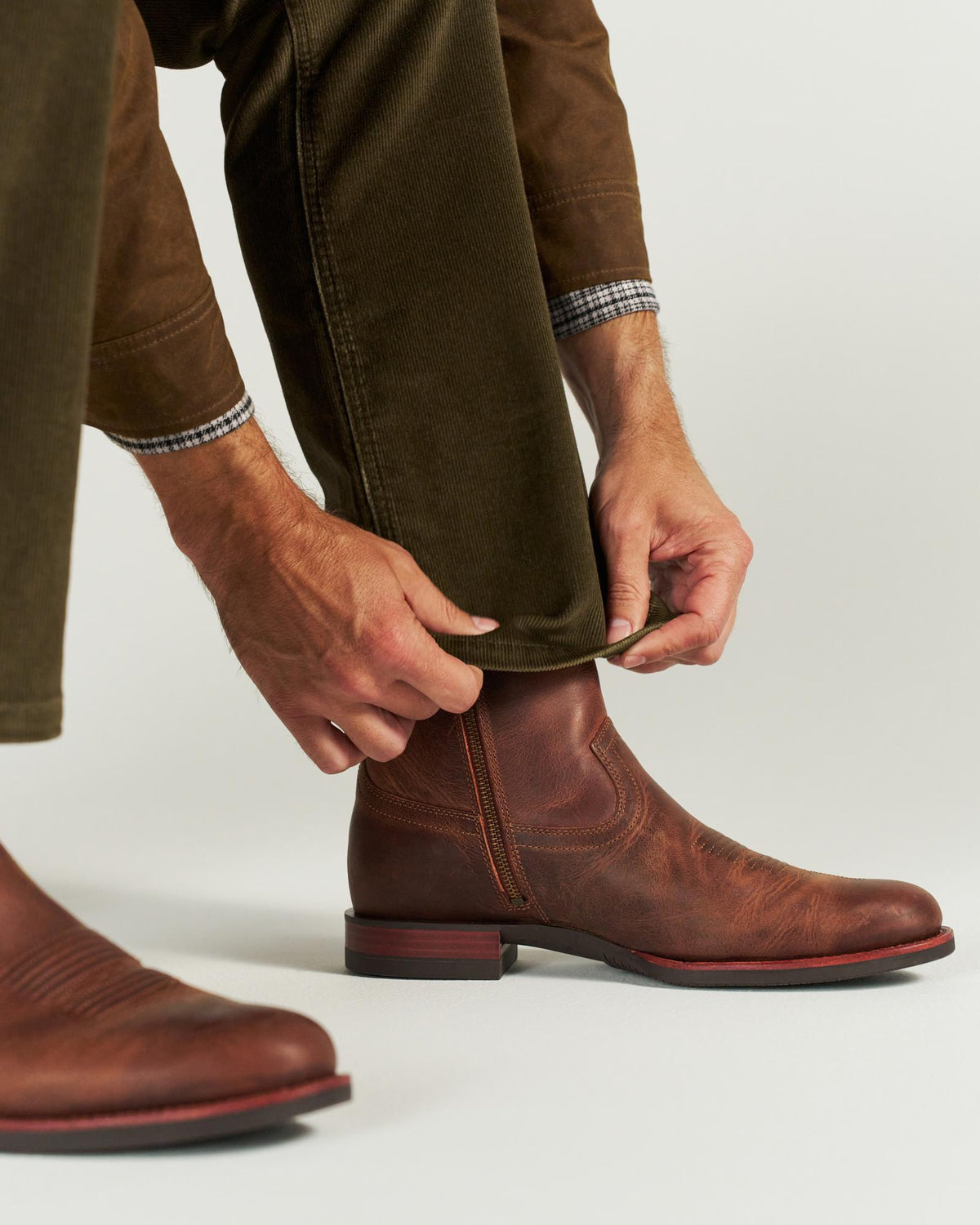 The Wayne in Maplewood Cowhide. A person wearing brown pants and plaid shirt cuffs tucks a pant leg into a Maplewood Cowhide brown leather boot with a red sole.