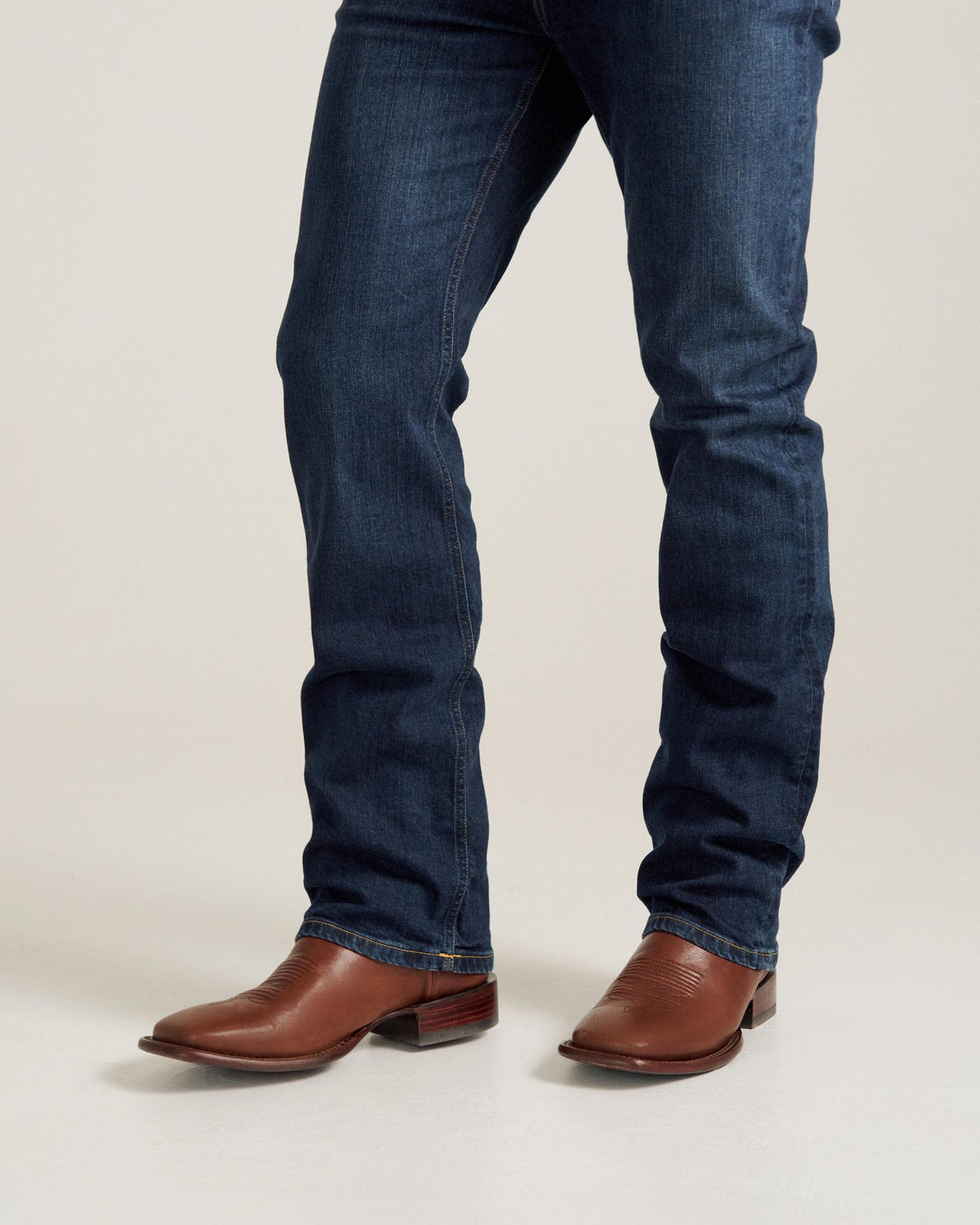 A person wearing blue jeans and brown leather western boots standing on a light-colored floor.