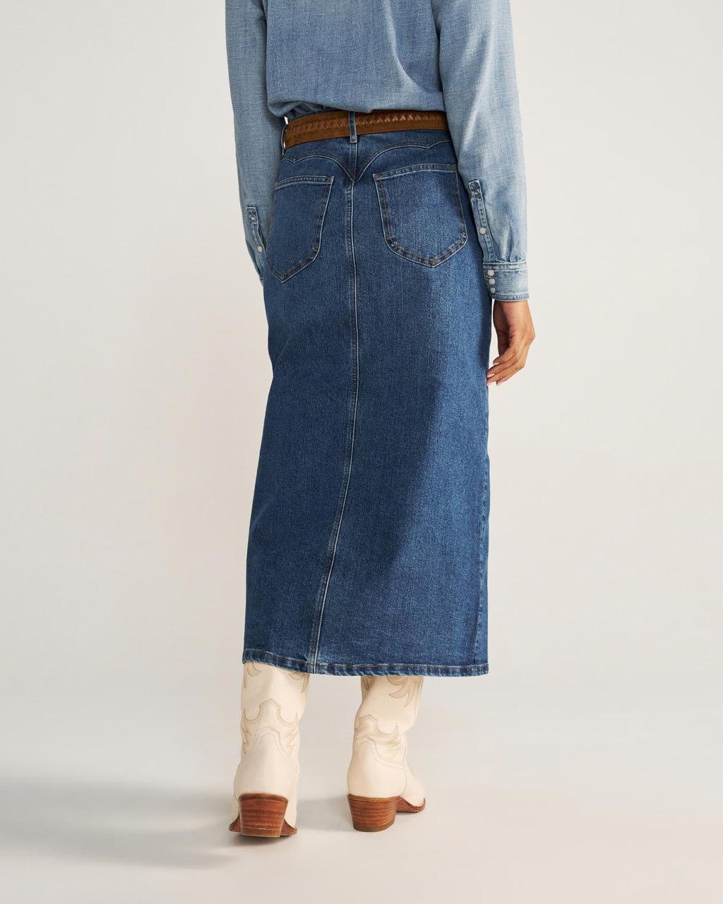 Person wearing a long denim skirt, brown belt, light blue denim shirt, and white cowboy boots, standing with their back to the camera.